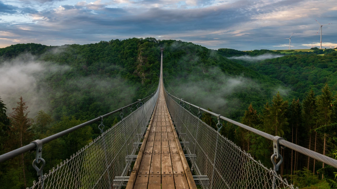 Blick auf eine lange Hängeseilbrücke, die sich über ein tiefes bewaldetes Tal erhebt, im Hintergrund bewölkter Himmel und Nebelschwaden im Tal