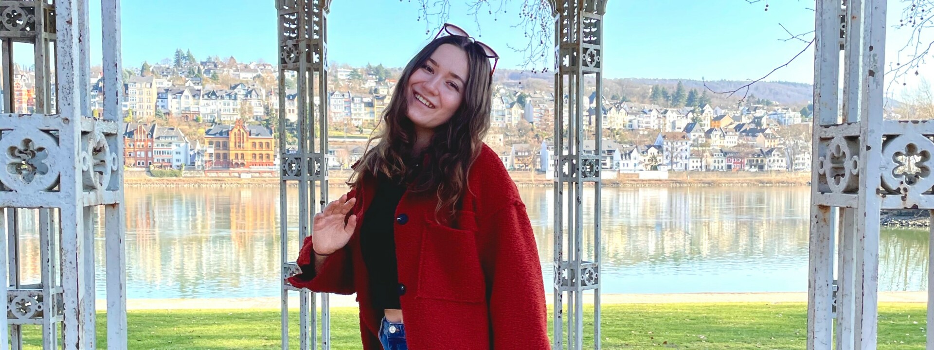 Frau mit rotem Mantel und Sonnenbrille steht im Augusta-Pavillon in den Rheinanlagen, im Hintergrund der Rhein und der Stadtteil Pfaffendorf. ©Kaya Reichert