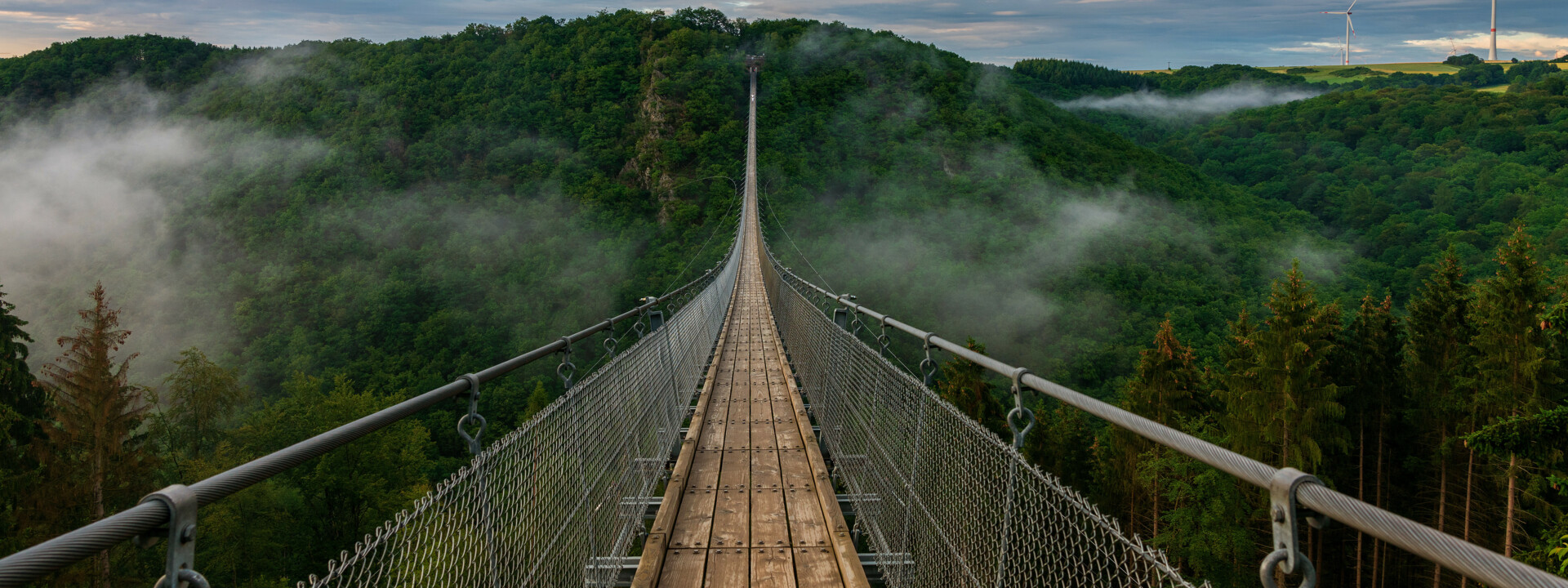 Blick auf eine lange Hängeseilbrücke, die sich über ein tiefes bewaldetes Tal erhebt, im Hintergrund bewölkter Himmel und Nebelschwaden im Tal  ©Bernhard - stock.adobe.com