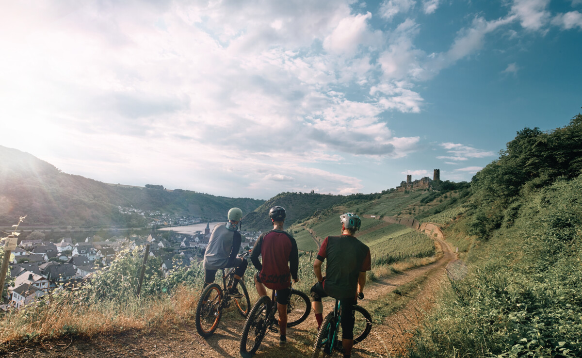 Freundesgruppe auf Mountainbikes auf einem Radweg neben der Mosel mit Burg Thurant im Hintergrund ©Philip Bruederle