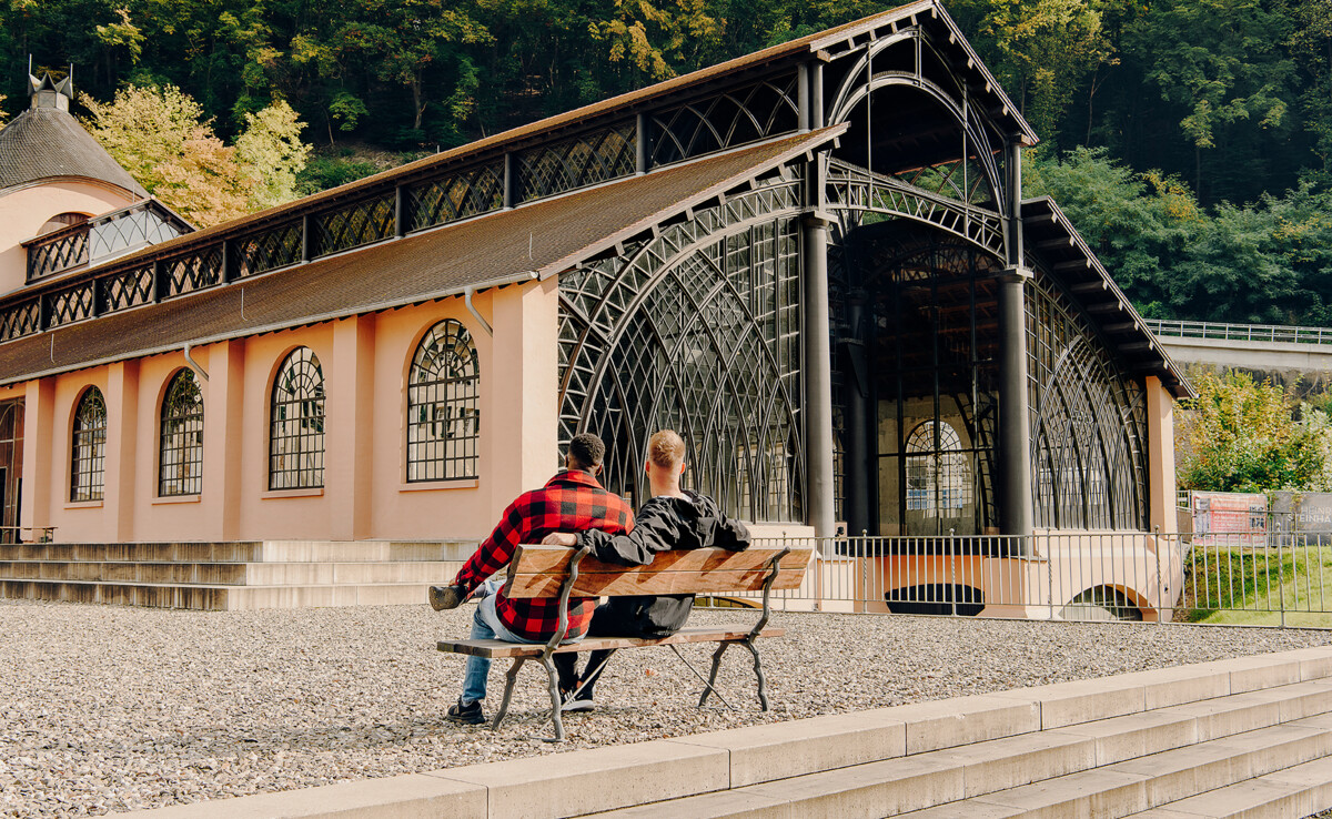 Zwei Männer von hinten sitzen auf einer Bank und blicken auf die Sayner Hütte ©Koblenz-Touristik GmbH, Philip Bruederle