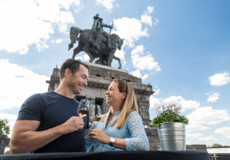 Pärchen steht mit Weinglas in der Hand vor dem Kaiser Wilhelm Denkmal am deutschen Eck und lachen sich gegenseitig an © Koblenz-Touristik GmbH, Dominik Ketz