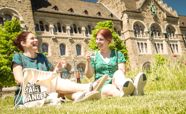 Zwei Mädels sitzen auf der Wiese vor dem preußischen Regierungsgebäude mit einem Glas Wein in der Hand und lachen. Der WeinStadtWander-Beutel steht vor ihnen auf der Wiese. ©Koblenz-Touristik GmbH, Johannes Bruchhof