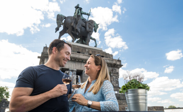 Couple standing in front of the Kaiser Wilhelm monument at the Deutsches Eck with wine glasses in their hands and laughing at each other ©Koblenz-Touristik GmbH, Dominik Ketz